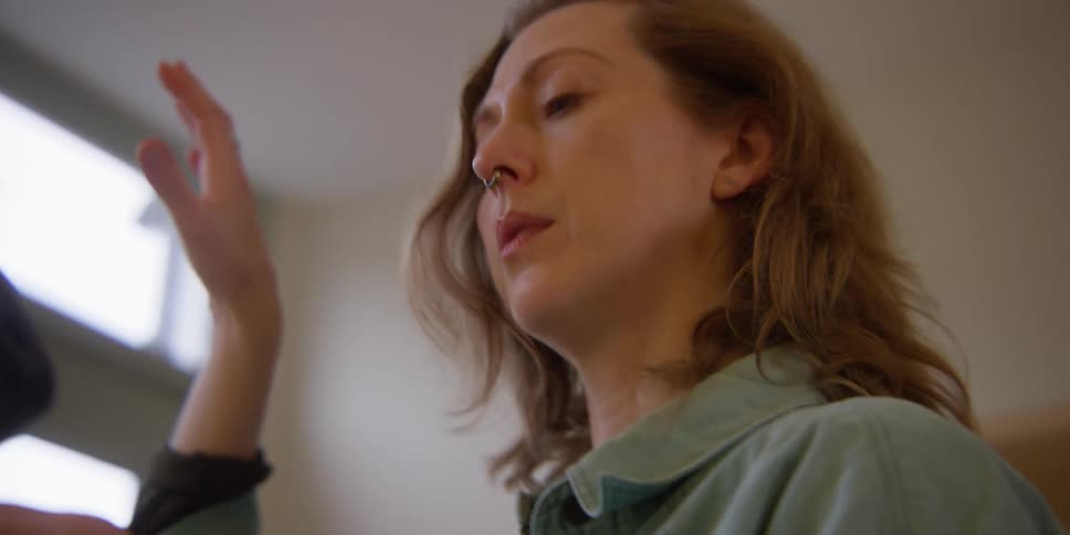 A red-headed female dance instructor with a septum ring reviews the recording of herself she just made on her smartphone, practices the dance moves with her hands, and then shakes her head and grimaces in mild frustration.