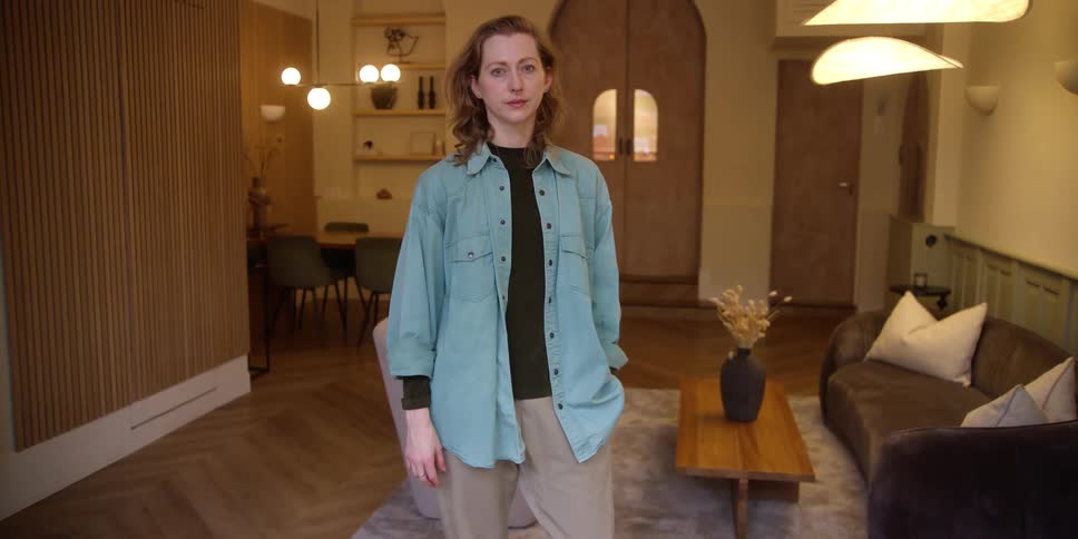 A white woman with red hair, fair skin, and a septum nose ring stands with a slight smile and her hand in her pocket in a mid-century modern style living room.