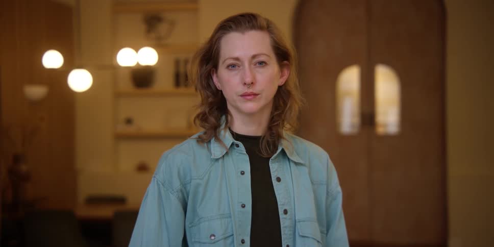 A white, red-headed woman with a septum ring standing in a mid-century style room looks at the camera without expression and then adjusts her head and smiles