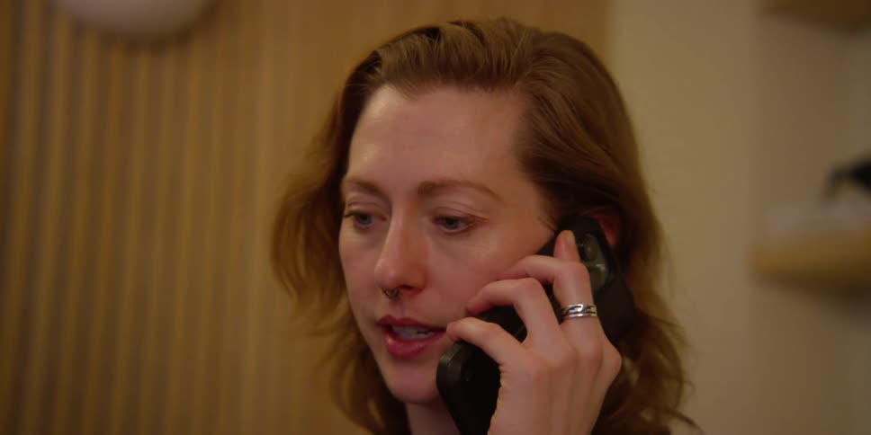 Close up of a fair-skinned woman with light eyes, red hair, and a nose ring who is indoors talking expressively on her cell phone before hanging up.