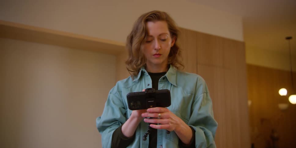 A red-headed Caucasian woman picks up her phone on a tripod and smiles and nods while viewing what she just recorded.