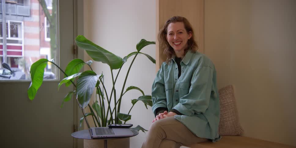 A fair woman with auburn hair smiles happily and sits on a bench near a window with her laptop on a table in front of her.