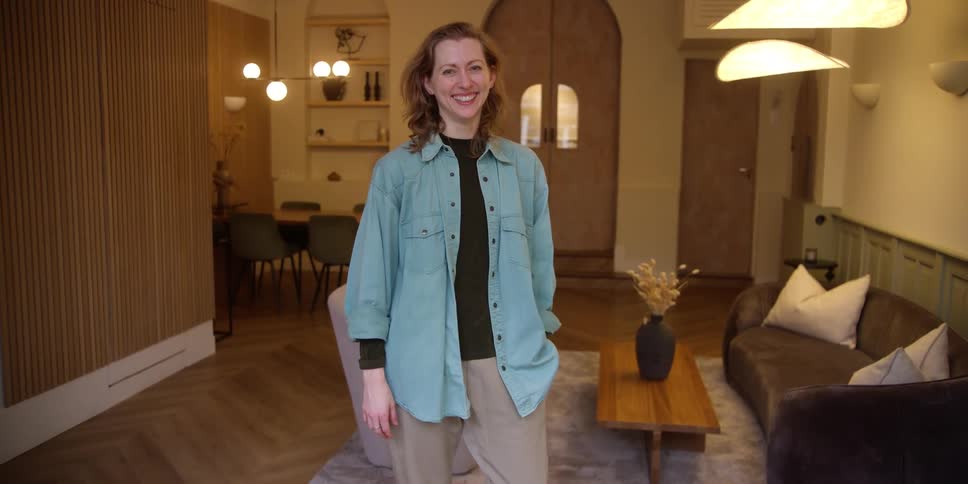 A ginger-haired white woman standing inside a retro living room and giving a beautiful smile with her hand in her pocket.