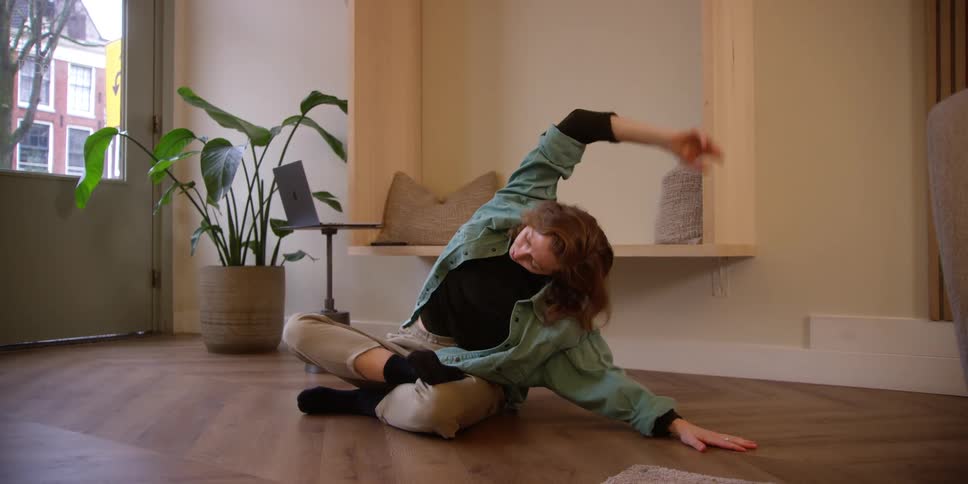An adult red-headed woman stretching her legs and arms while sitting on the floor of an apartment with views to the city outside.