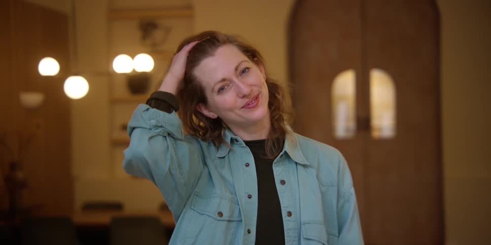 A red-headed woman with a septum ring standing in a mid-century style room looks at the camera with a laughing smile and pushes her hair back with her hand.