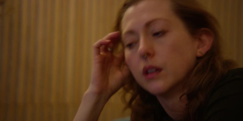 A fair-skinned woman with red hair and a septum ring rolls her eyes and gestures in a frustated manner as she looks down at her laptop.