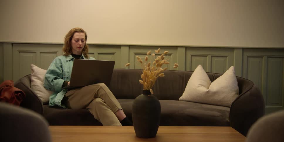 An indoor setting with a woman sitting on a dark retro sofa working on a laptop as the camera moves forward and then back.