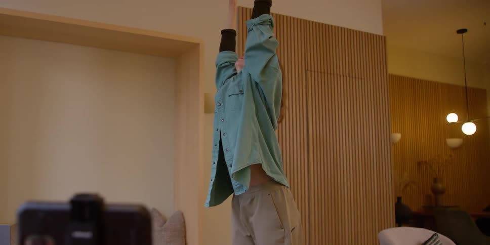 A red-headed woman films herself practicing contemporary dance moves in a room with a smartphone on a tripod.