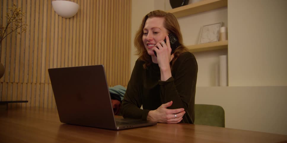 A fair, red-headed woman is sitting in a mid-century dining area and typing on her laptop when she stops to answer a phone call on her cell phone with a big smile.