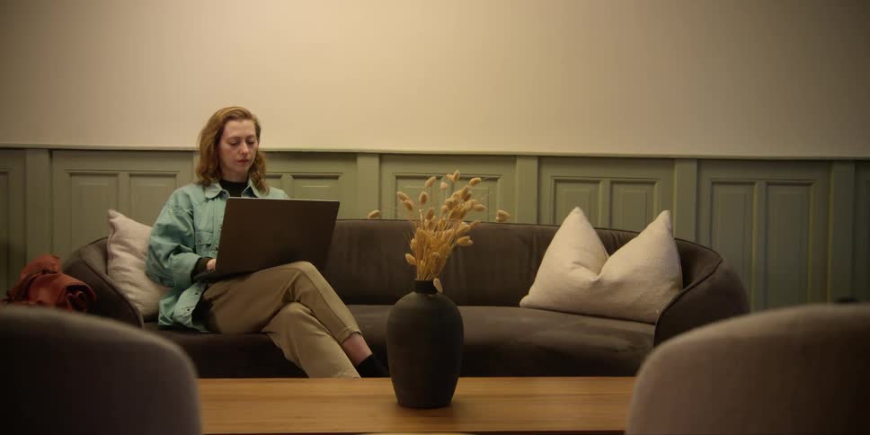 A fair red-headed woman is sitting on a sofa while using a laptop in a mid-century modern living area.