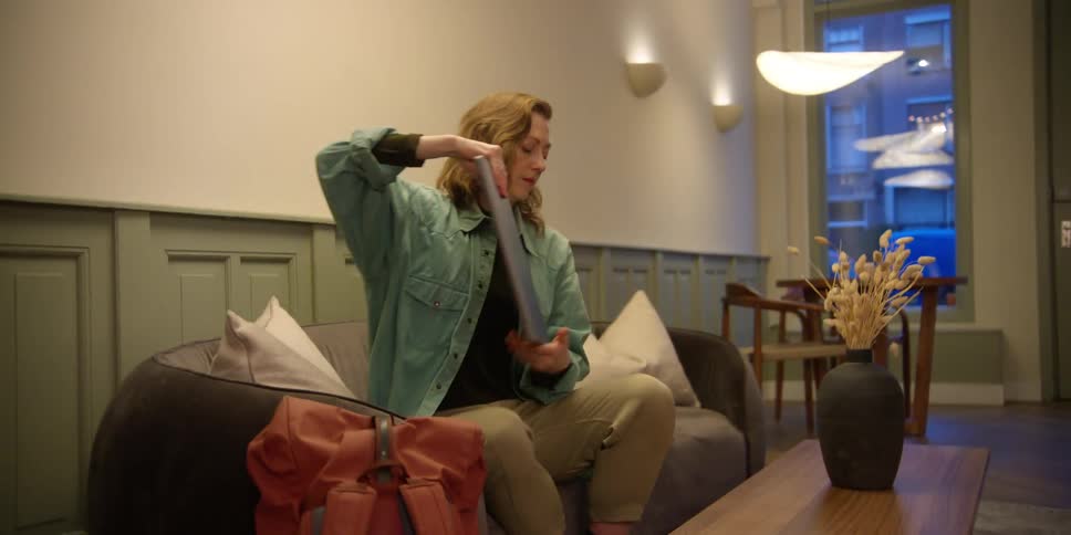 A young, red-headed woman sitting on a couch takes a laptop from a backpack and opens it on a coffee table