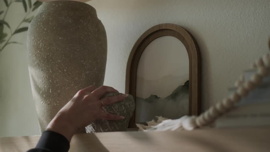 A person arranges a painting and a stone bowl on a wooden shelf