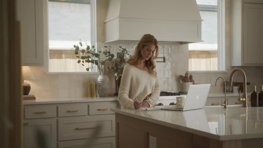 A woman in a kitchen copies with pen and paper something shes reading from a laptop