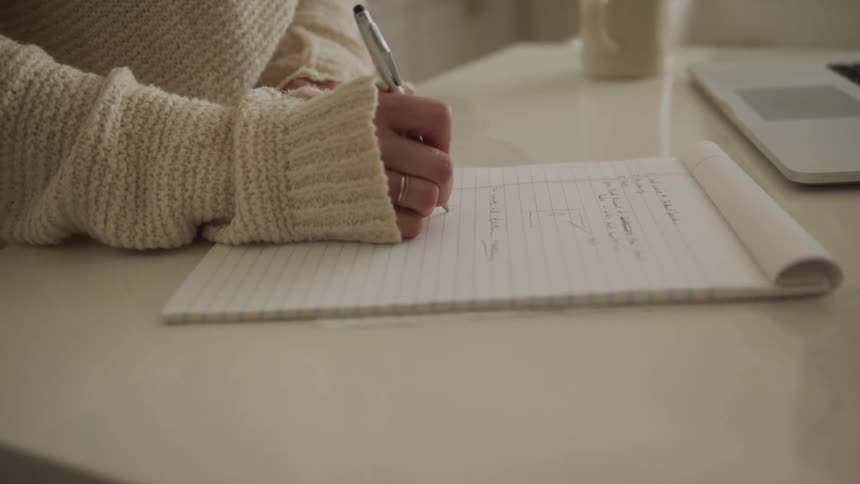A white woman uses a laptop and then writes on a pad of paper