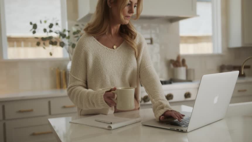 Woman in the kitchen standing drinking a cup of coffee typing on her laptop and writing in a notebook