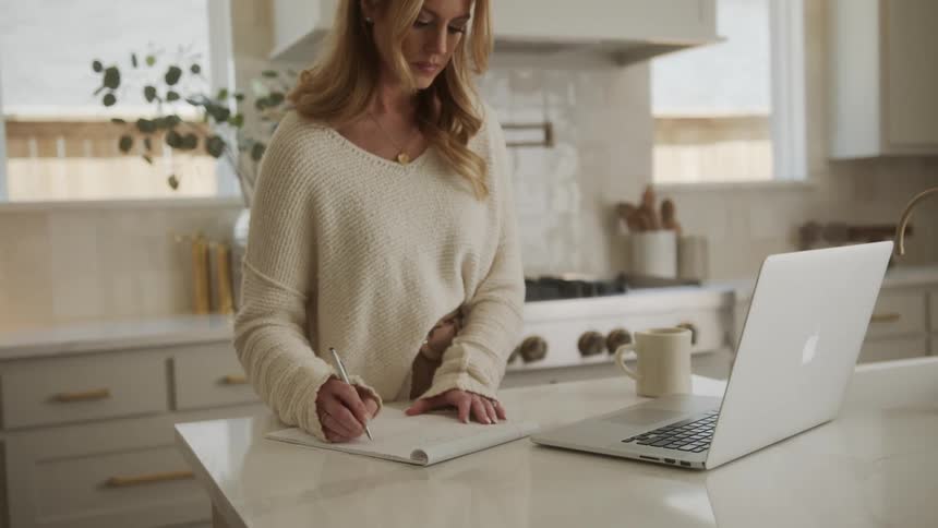Woman in the kitchen working on her laptop and writing in a notebook in a concentrated manner
