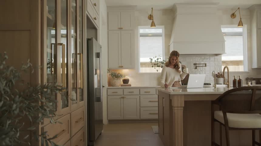 Woman in the kitchen drinking a cup of coffee and standing in front of the counter writing in a notebook with her laptop beside her