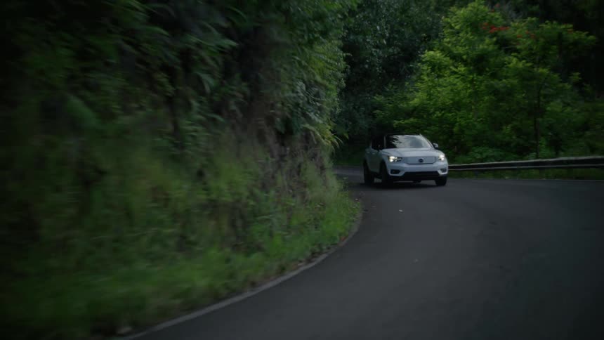 A white automobile travels on a curved road out of the dark forest