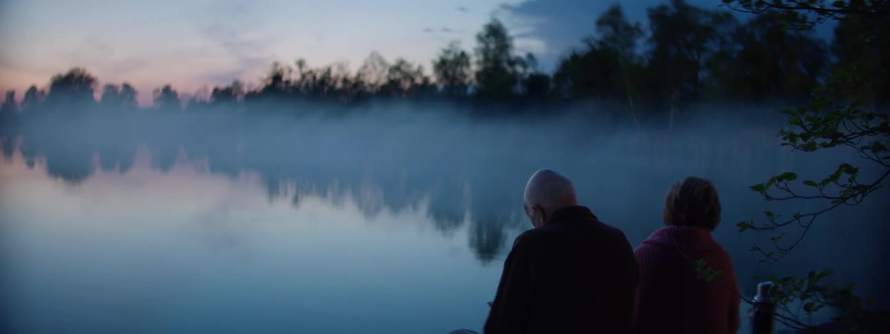 Two older people sit next to a large lake