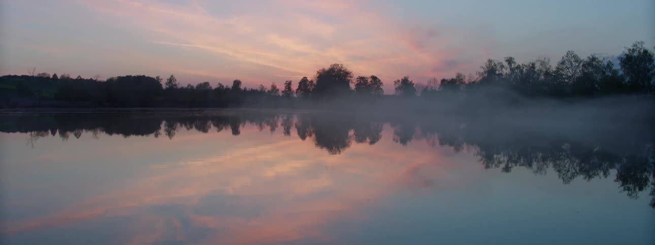 A highly reflective lake is lined with trees under a sky filled with wispy clouds