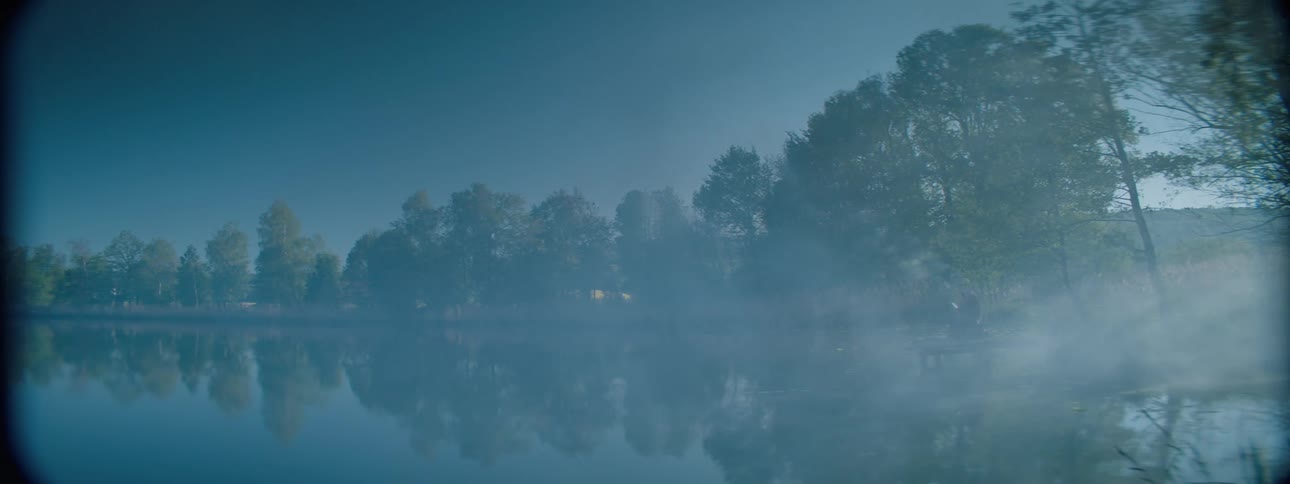 A lake with many trees around it is reflecting them on the surface of the water and there is a dense fog over the region