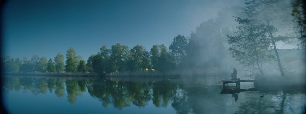 Person on pier over reflective water is silhouetted by fog