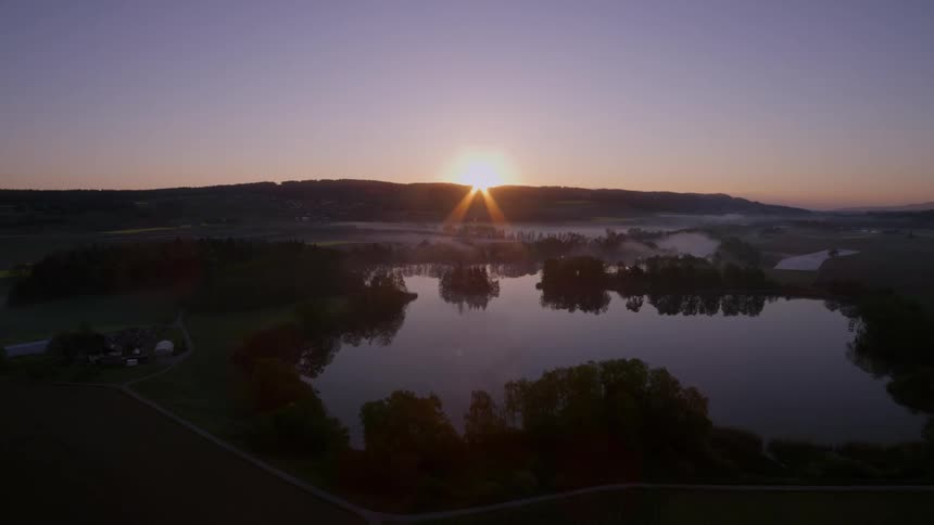 An aerial view of a lake that is reflecting the trees that are around it and on the horizon the sun that is rising one morning