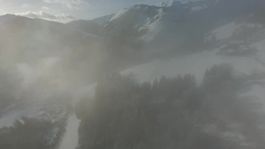 An aerial view descending from a snowy mountain and pine trees and there are clouds on the way