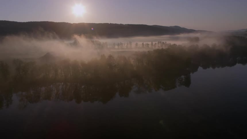 A lake reflecting several trees that are on the shore and on top of them there is a mysterious fog and there is a strong sunlight