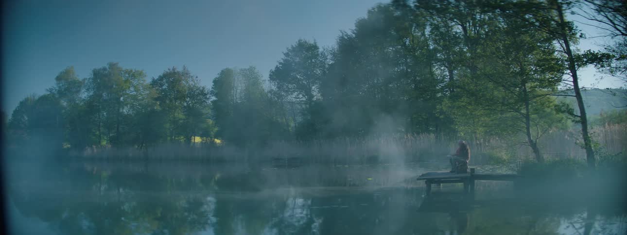 A person sitting on a deck looking at a lake that is filled with fog on the surface