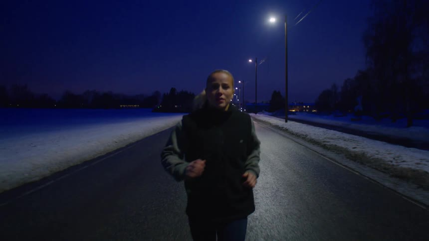 A young blonde woman jogs down a road on a winter night