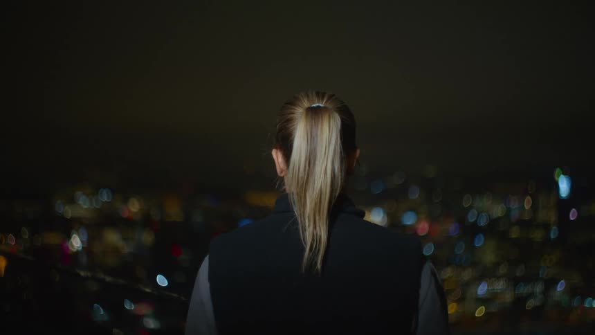 A woman wearing a ponytail and a black vest gazes at the city skyline illuminated by the night