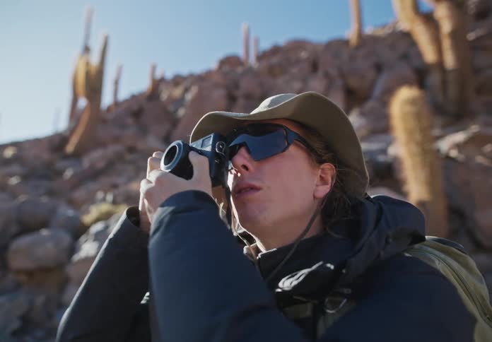 Portrait of a woman with a digital camera adjusting while standing in an outdoor environment of desert vegetation with rocks around