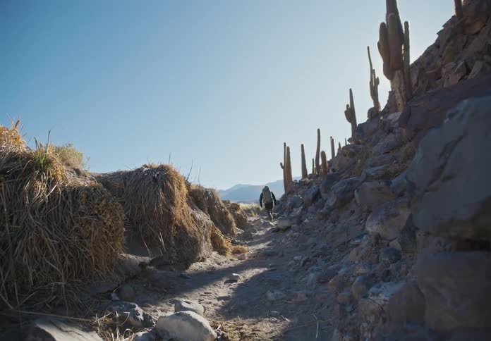 A hiker with a backpack and wearing pants navigates through a landscape of hay and rocky mountain slopes