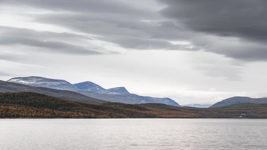 A big lake with some ripples moving around and on the land there is a forest full of trees and in the sky there are some gray clouds moving