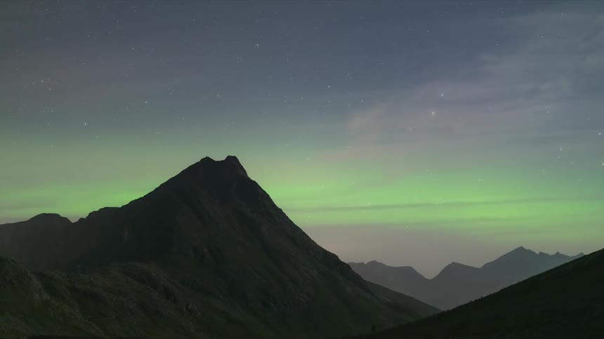 Wispy clouds pass through green and purple night sky over mountain range