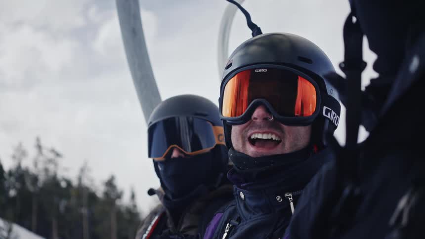 Two men ride a ski lift one man smiles as he looks at the camera laughing