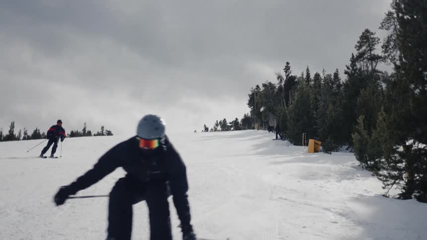 Multiple skiers ski down wide snowy slope between conifers on an overcast day