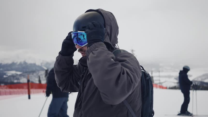A man is wearing a ski goggle on his helmet and he puts it on his eyes in a region with a lot of snow on the ground and some people with skis behind