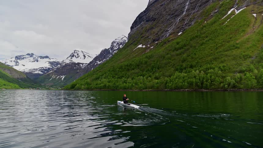 Man riding a canoe along a river surrounded by mountains with vegetation and snow