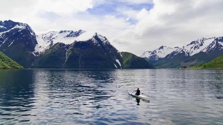A man rows a boat across a picturesque inlet in Norway surrounded by rugged terrain and snowy mountains