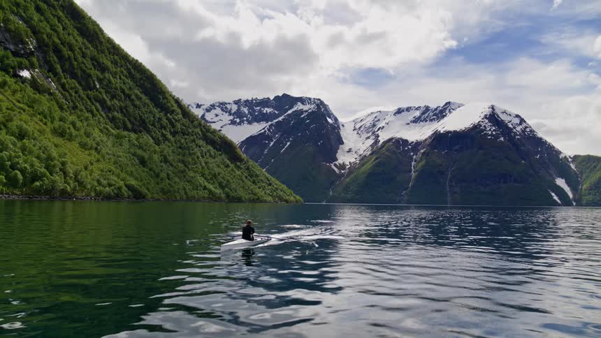 A man rows a boat on glassy water in a valley surrounded by mountainous terrain