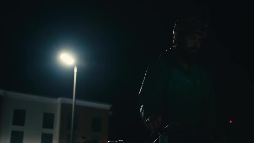 A cyclist walks away from a building with his bicycle at his side then stops and closes his eyes with a sad expression