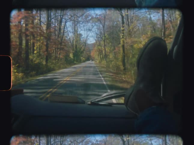 A car traveling down a treelined road with passengers feet resting on the dashboard