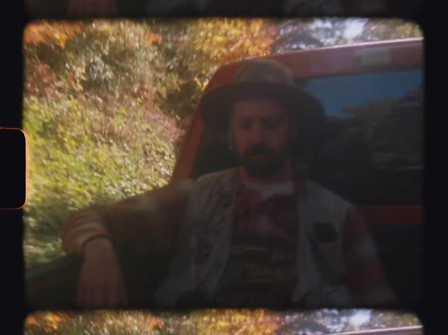 Two men sit in the back of a pickup truck driving through a forest