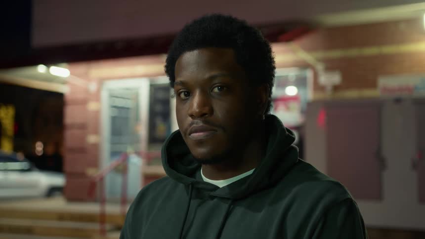 A black man in a hoodie poses outside of a building in its parking lot