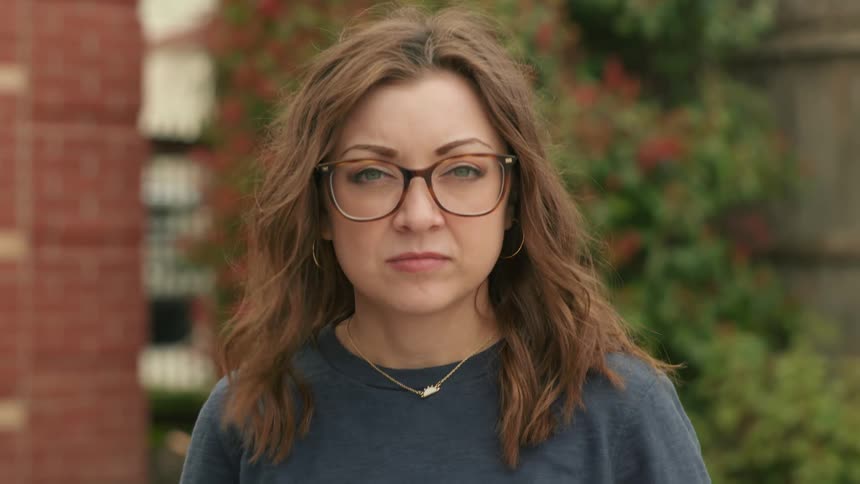 A stoic woman with glasses turns her head