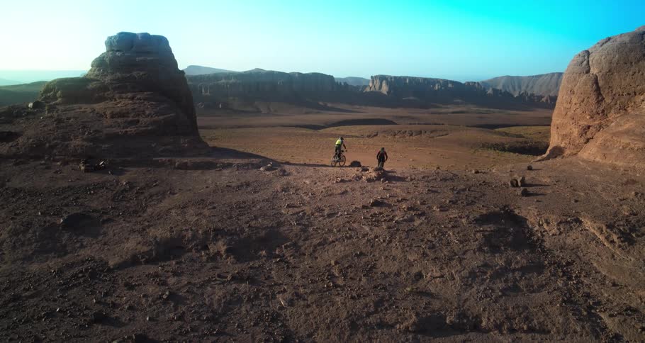 Two cyclists are riding on a rugged terrain with large rock formations under a clear sky