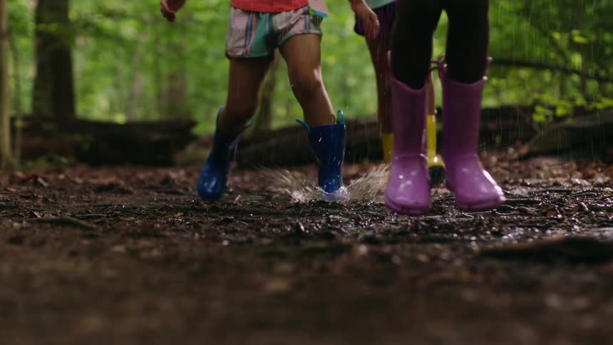 Legs of three girls with colorful boots jumping and walking on a ground in the forest on a day with rain
