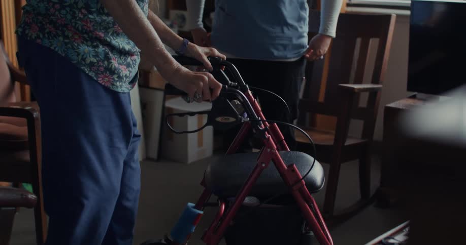 Elderly woman uses a walker to cross the room with a medical professional looking on